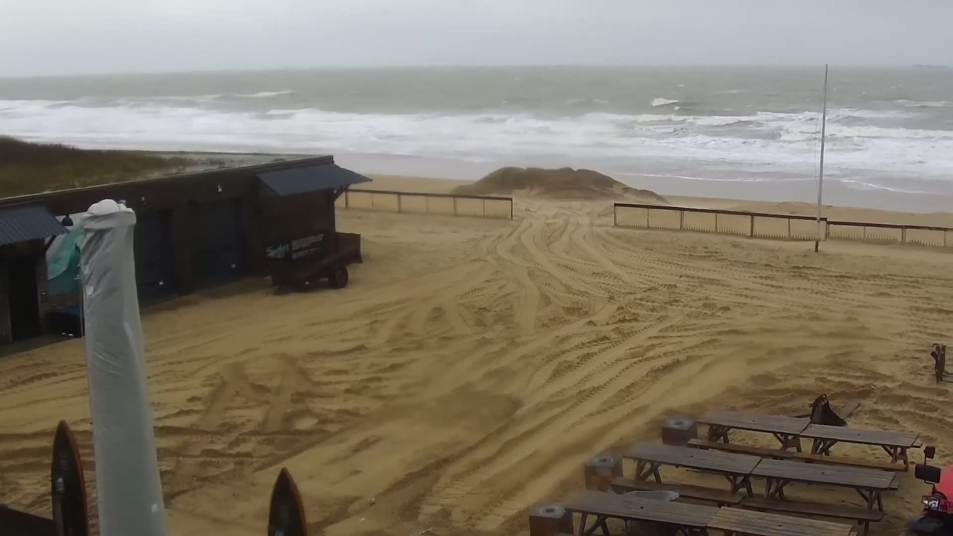 Two people stand on a wooden viewing platform overlooking a wide, sandy beach and a grey, wavy ocean under an overcast sky.