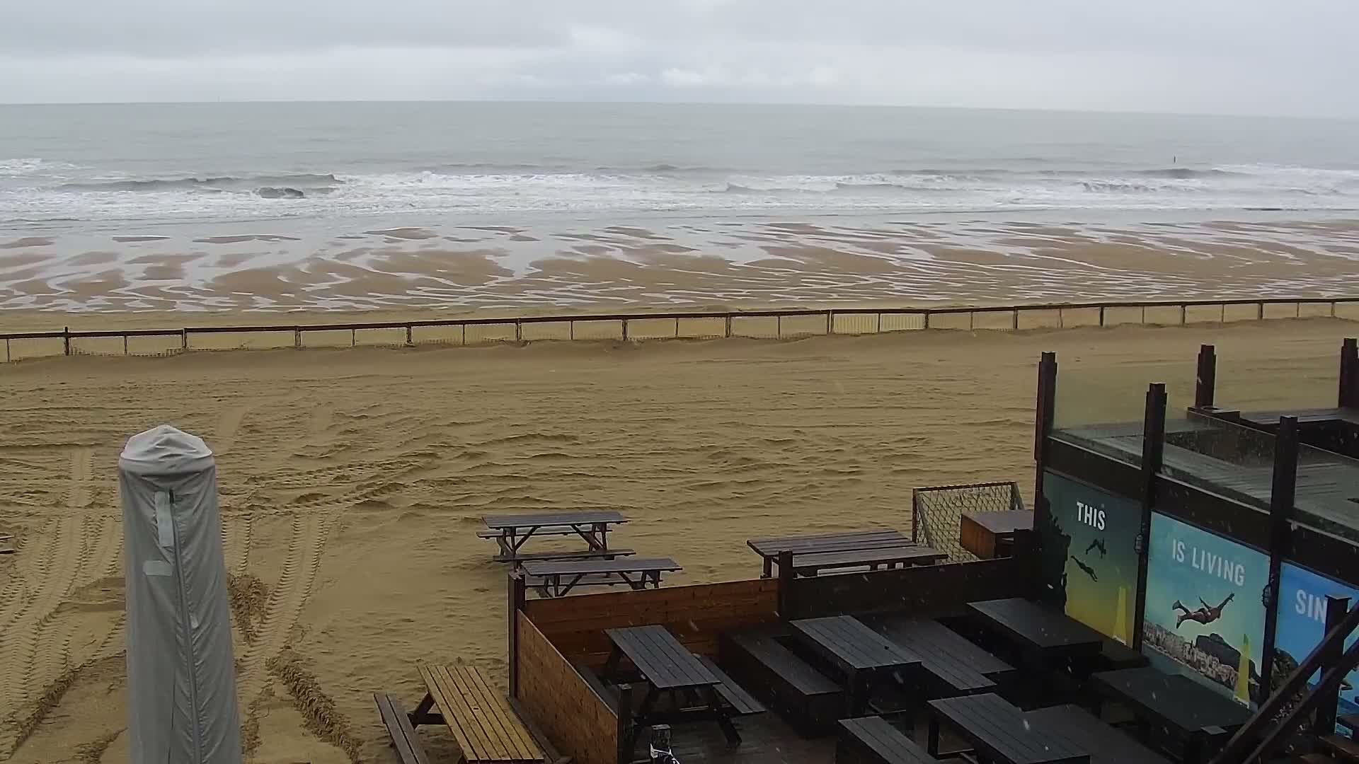 A wide, sandy beach with wet patterns from receding waves is separated by a fence from an outdoor seating area featuring numerous picnic tables and a covered umbrella, all under an overcast sky with light snow or sleet falling.