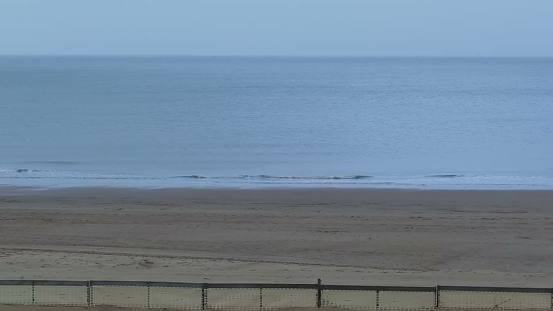 A sandy beach and a calm ocean with gentle waves under a soft, overcast, and hazy sky, with a light-colored fence visible at the bottom of the frame.