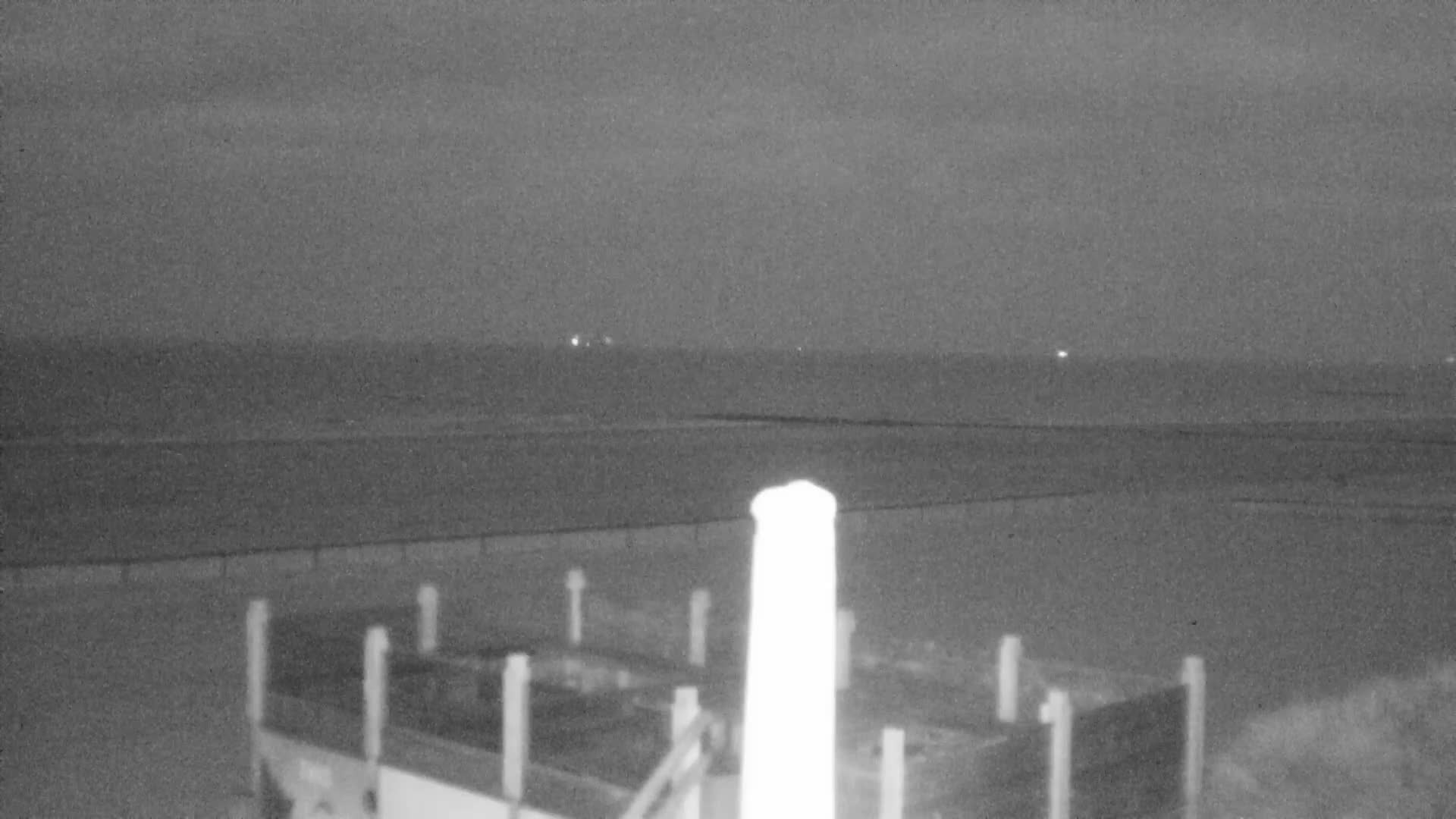 A wide sandy beach meets the calm ocean under an overcast sky, with a tall white pole in the center foreground and dark rocks visible on the right.