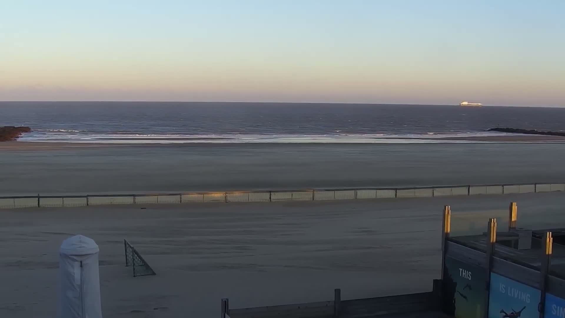 A wide, empty sandy beach stretches towards a calm ocean with gentle waves under a clear, pastel-hued sky, featuring a distant ship, rock jetties, a covered beach umbrella, and a small soccer goal, indicating a clear and peaceful day.