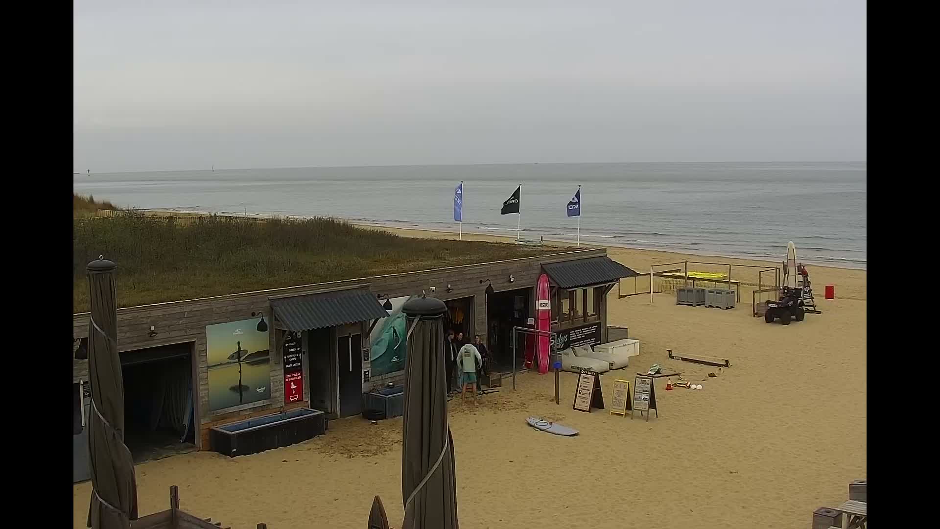 A wide, empty sandy beach stretches towards a calm ocean with gentle waves under a clear, pastel-hued sky, featuring a distant ship, rock jetties, a covered beach umbrella, and a small soccer goal, indicating a clear and peaceful day.