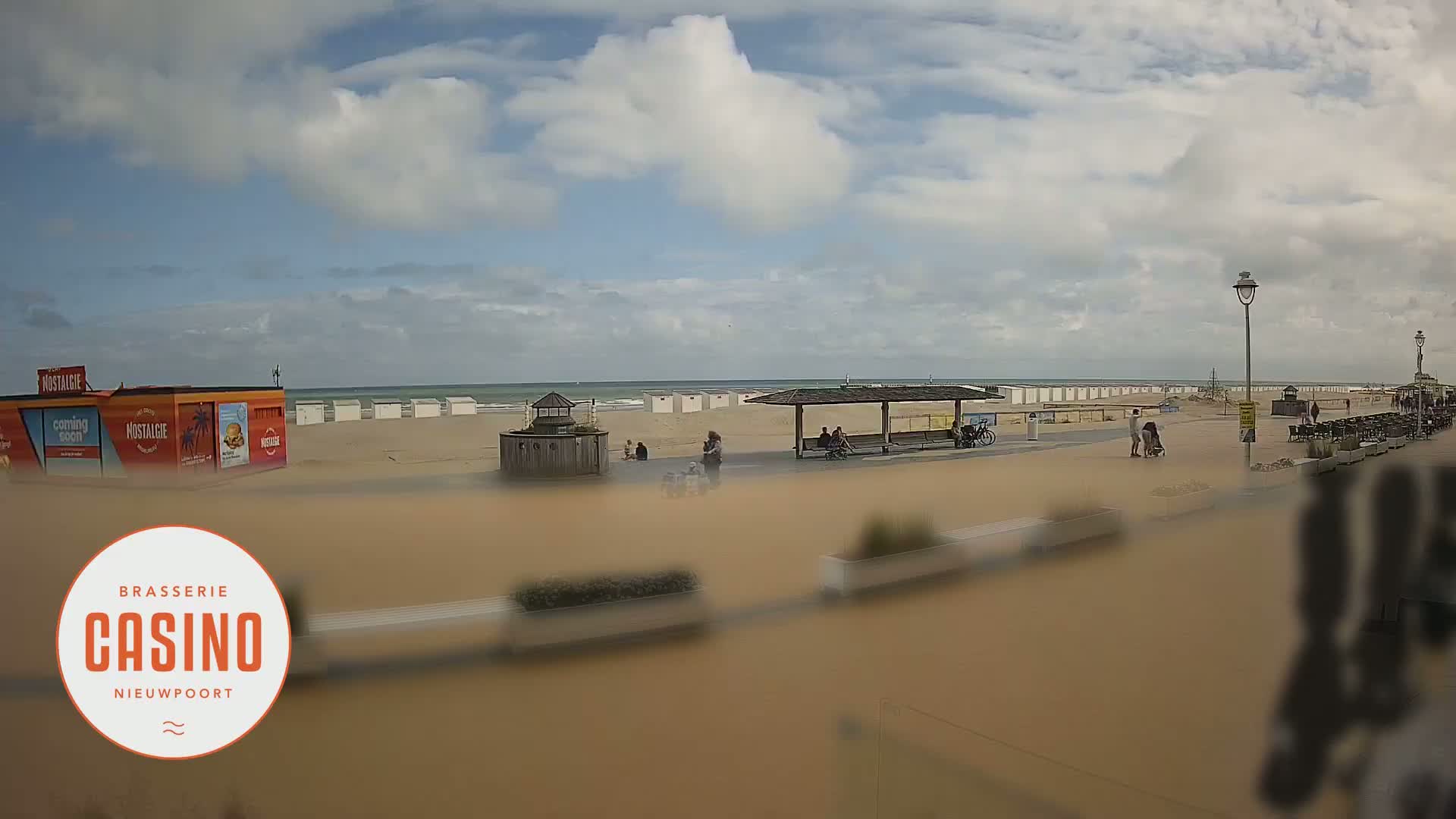 A sandy beach with people and beach huts under a partly cloudy sky, with a calm ocean in the background.