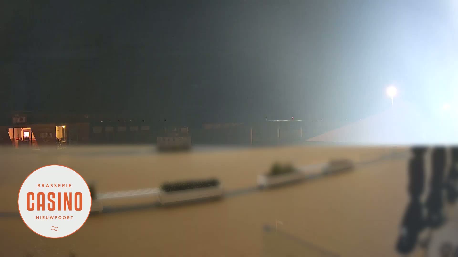 A foggy night scene shows a brightly lit area with a building in the background.