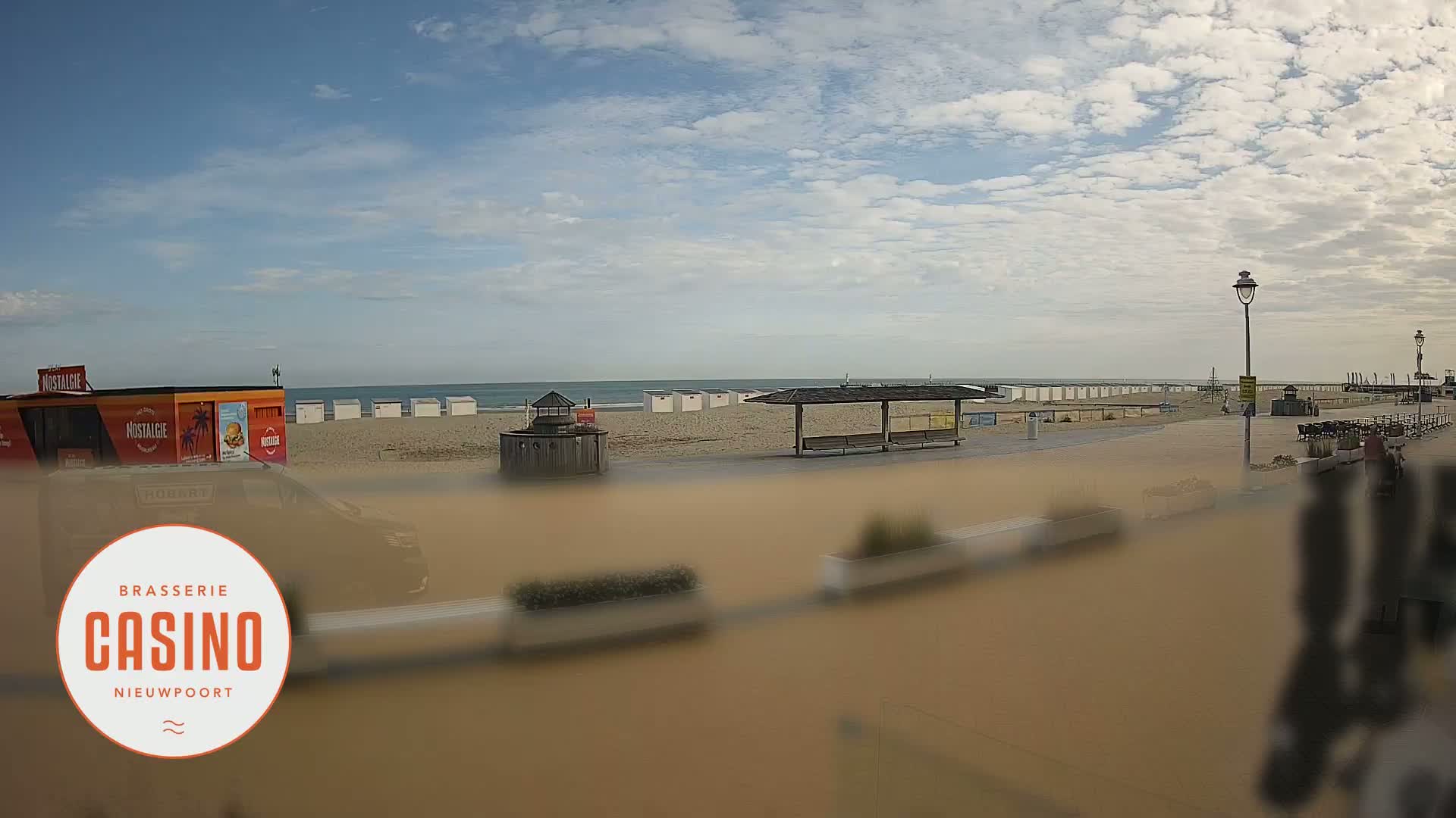 A partly cloudy day at a beach shows white beach huts, a boardwalk, and a calm ocean.