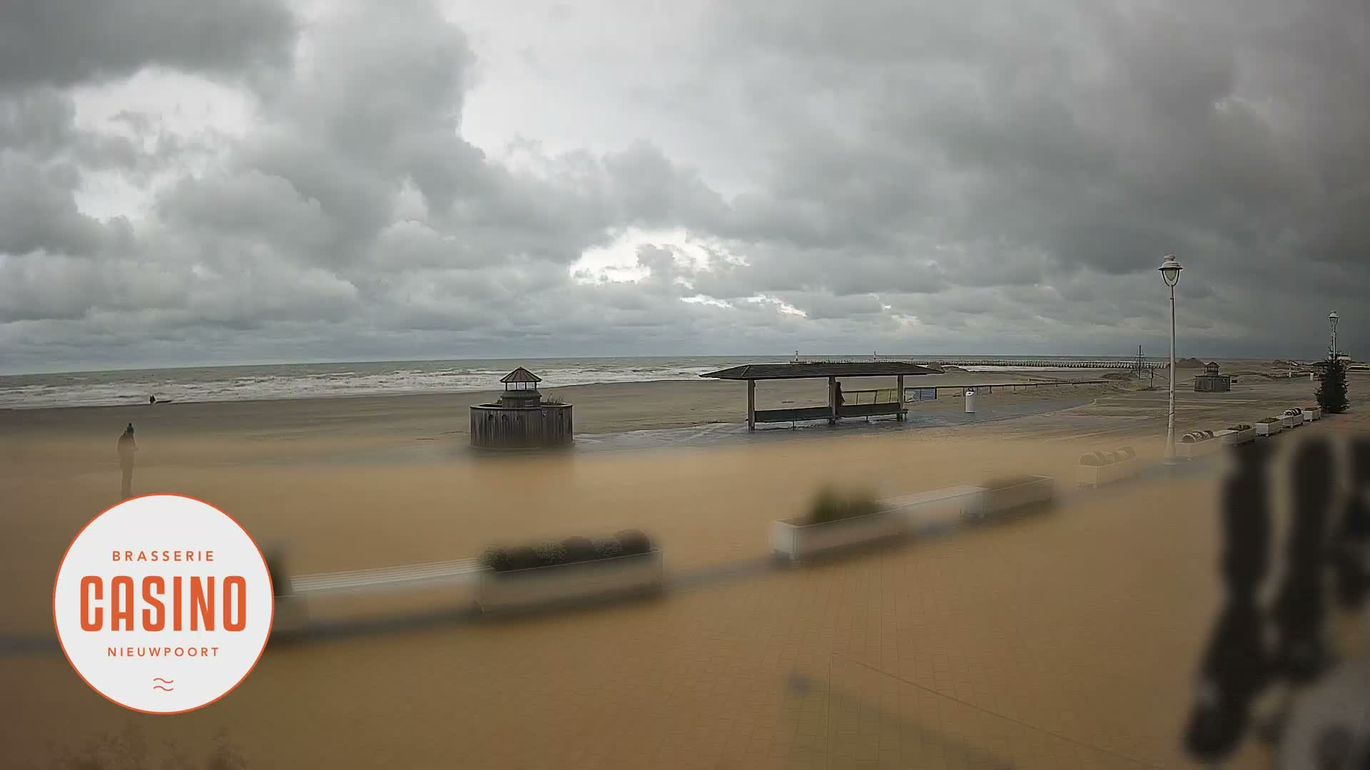 A wide, sandy beach and promenade are visible under a heavily overcast sky with choppy ocean waves, featuring several benches, streetlights, and a few small structures, with distant figures scattered across the sand.
