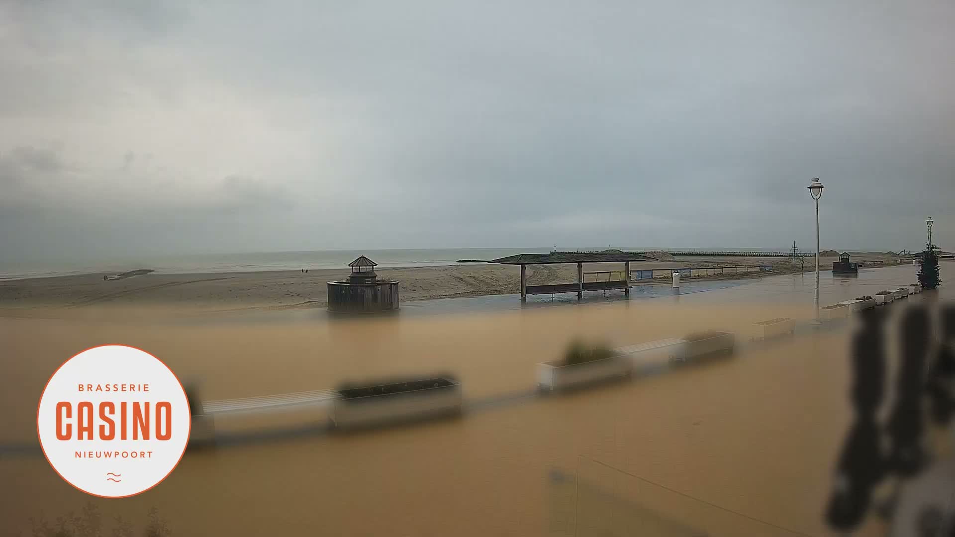 On an overcast day, a wide, wet promenade stretches along a sandy beach with the sea beyond, featuring a small wooden hut, a covered seating area, and distant figures under a grey sky.