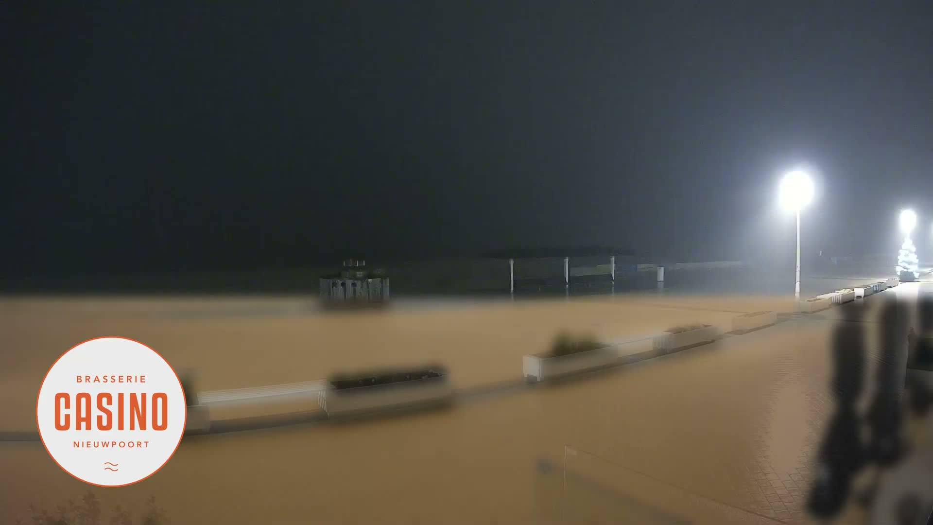 On an overcast day, a wide, wet promenade stretches along a sandy beach with the sea beyond, featuring a small wooden hut, a covered seating area, and distant figures under a grey sky.