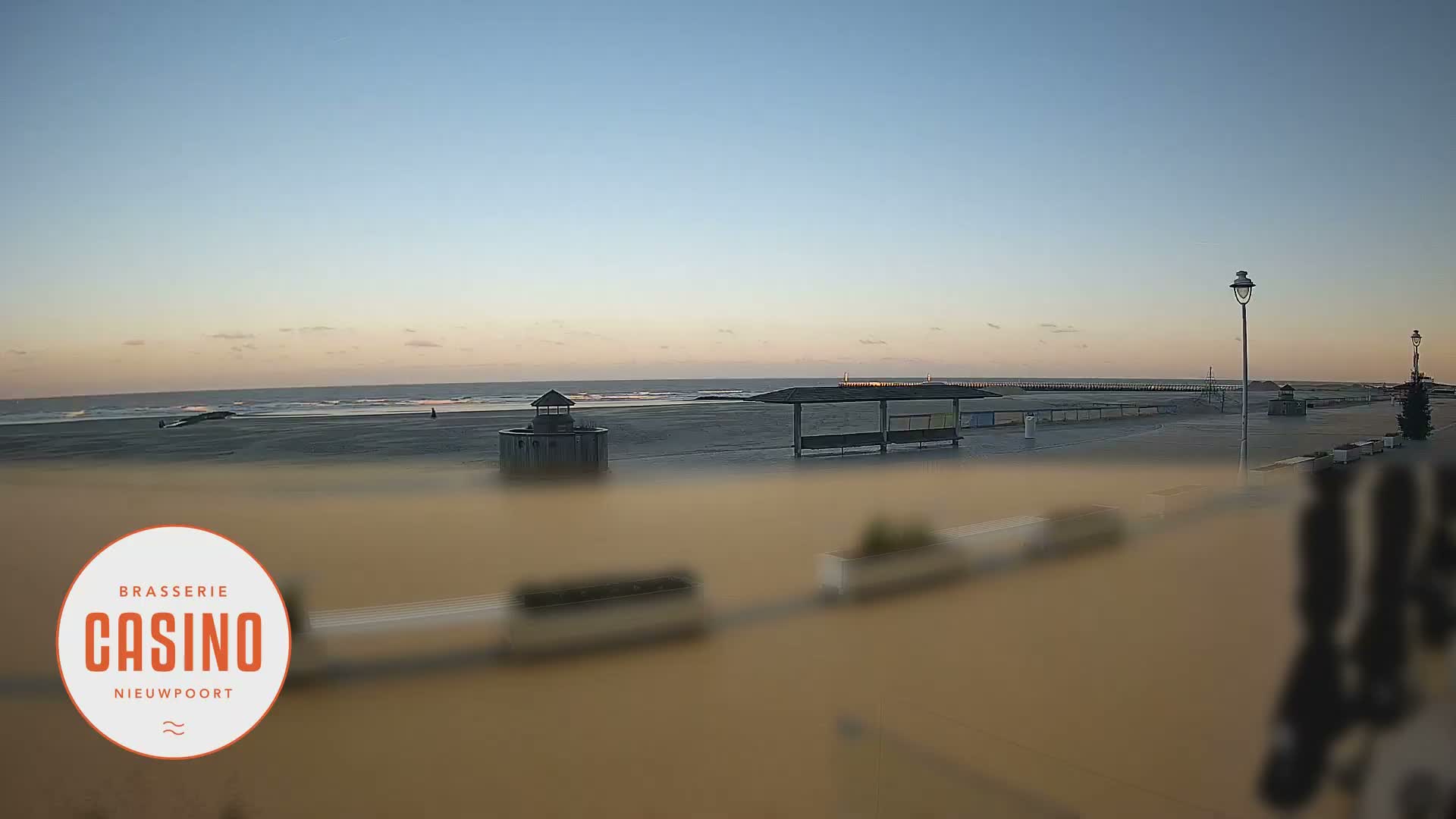 A tranquil beach and promenade are visible at dawn or dusk under a clear, calm sky with soft pastel hues, featuring gentle waves, a distant pier, a covered shelter, a small round building on the sand, and streetlights along the foreground walkway.