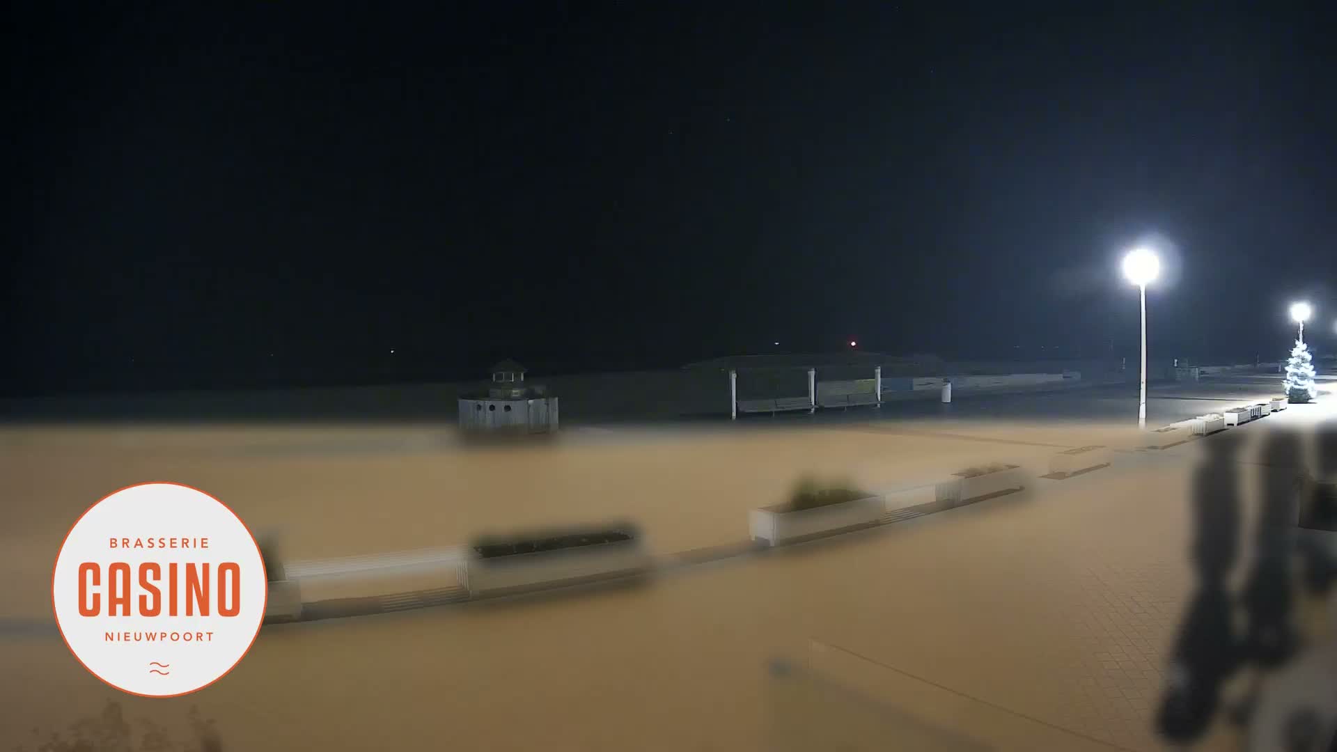 A tranquil beach and promenade are visible at dawn or dusk under a clear, calm sky with soft pastel hues, featuring gentle waves, a distant pier, a covered shelter, a small round building on the sand, and streetlights along the foreground walkway.