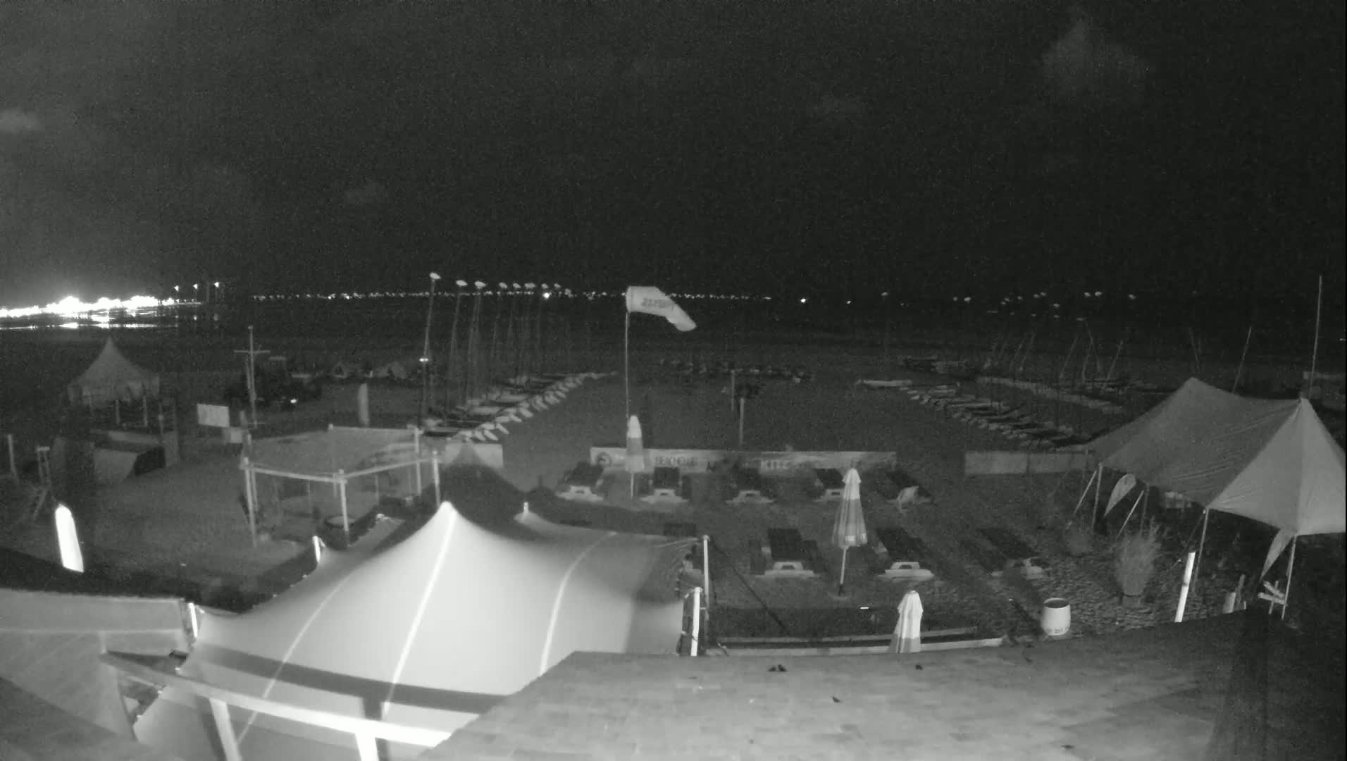 A nighttime beach scene shows several tents and numerous sailboats lined up on the sand, with a calm body of water visible in the background under a partly cloudy sky.