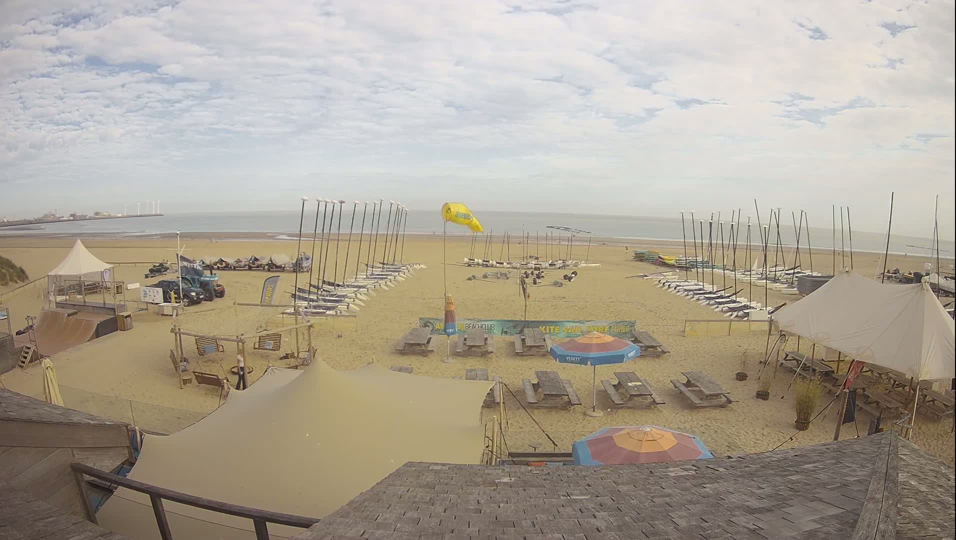An overhead view shows a sandy beach with numerous sailboats and equipment lined up, picnic tables, tents, and a calm ocean under a partly cloudy sky.