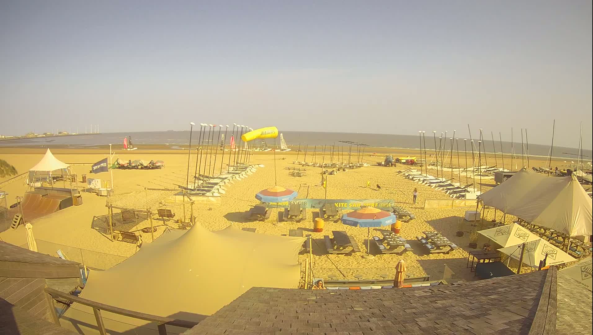 A sandy beach with numerous sailboats and beach umbrellas under a clear, sunny sky.