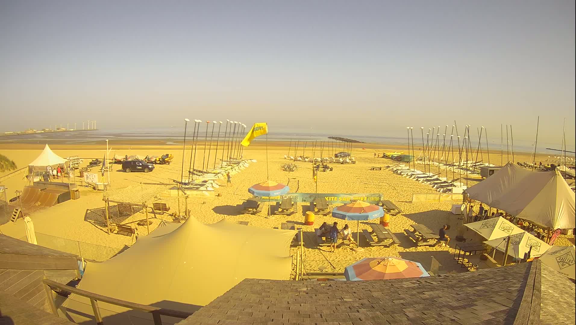A sandy beach on a sunny day with numerous sailboats and windsurfing equipment lined up, along with several tents, picnic tables with umbrellas, and people gathered in a beachside area.