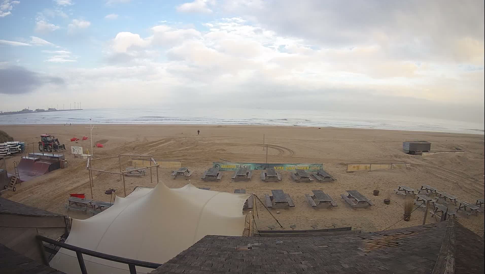 An aerial view reveals a wide, sandy beach equipped with numerous picnic tables, a large tent, a tractor, and watersports gear, all under a partly cloudy sky, leading to a calm ocean with distant wind turbines and industrial structures.