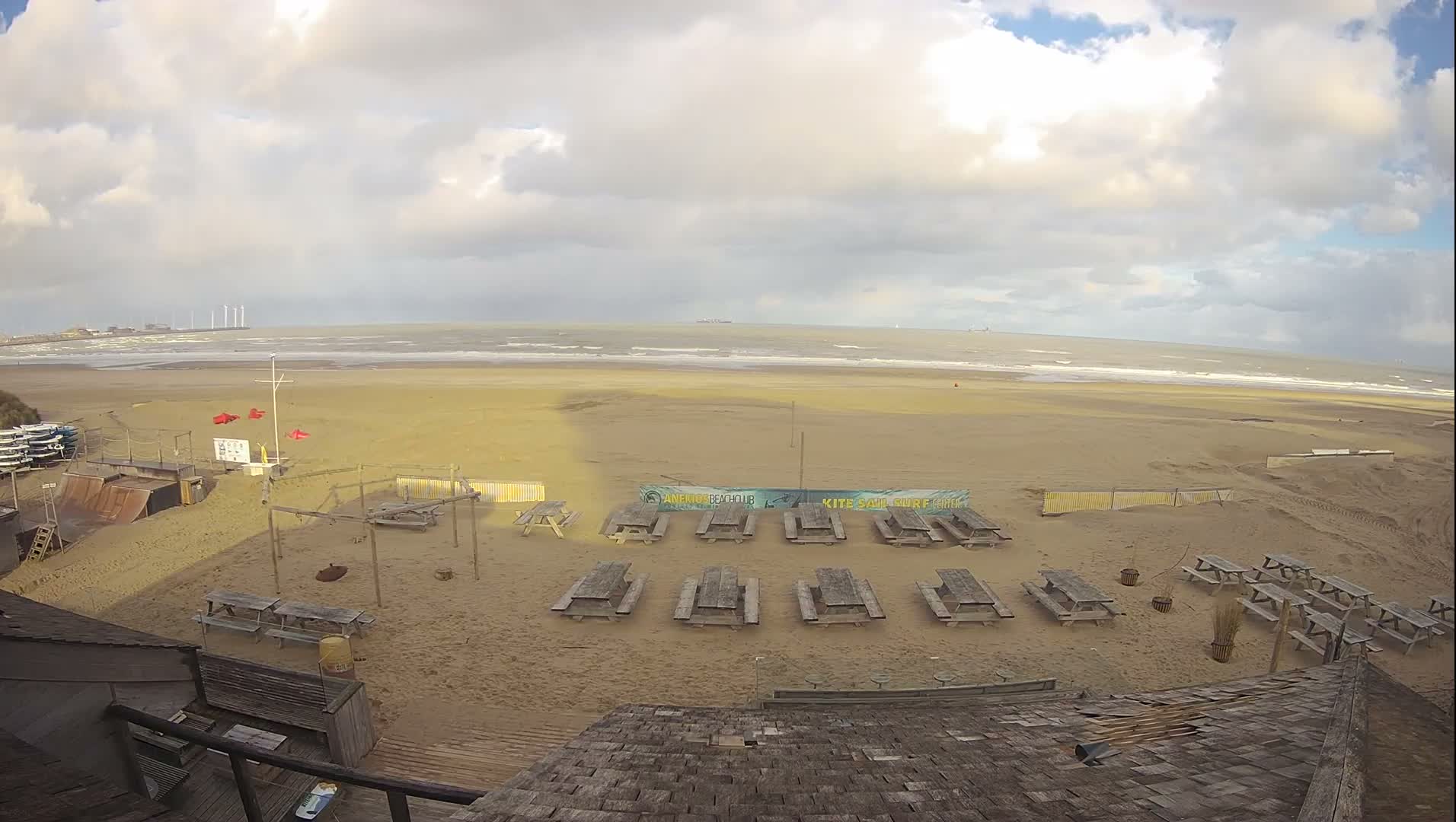 An aerial view reveals a wide, sandy beach equipped with numerous picnic tables, a large tent, a tractor, and watersports gear, all under a partly cloudy sky, leading to a calm ocean with distant wind turbines and industrial structures.