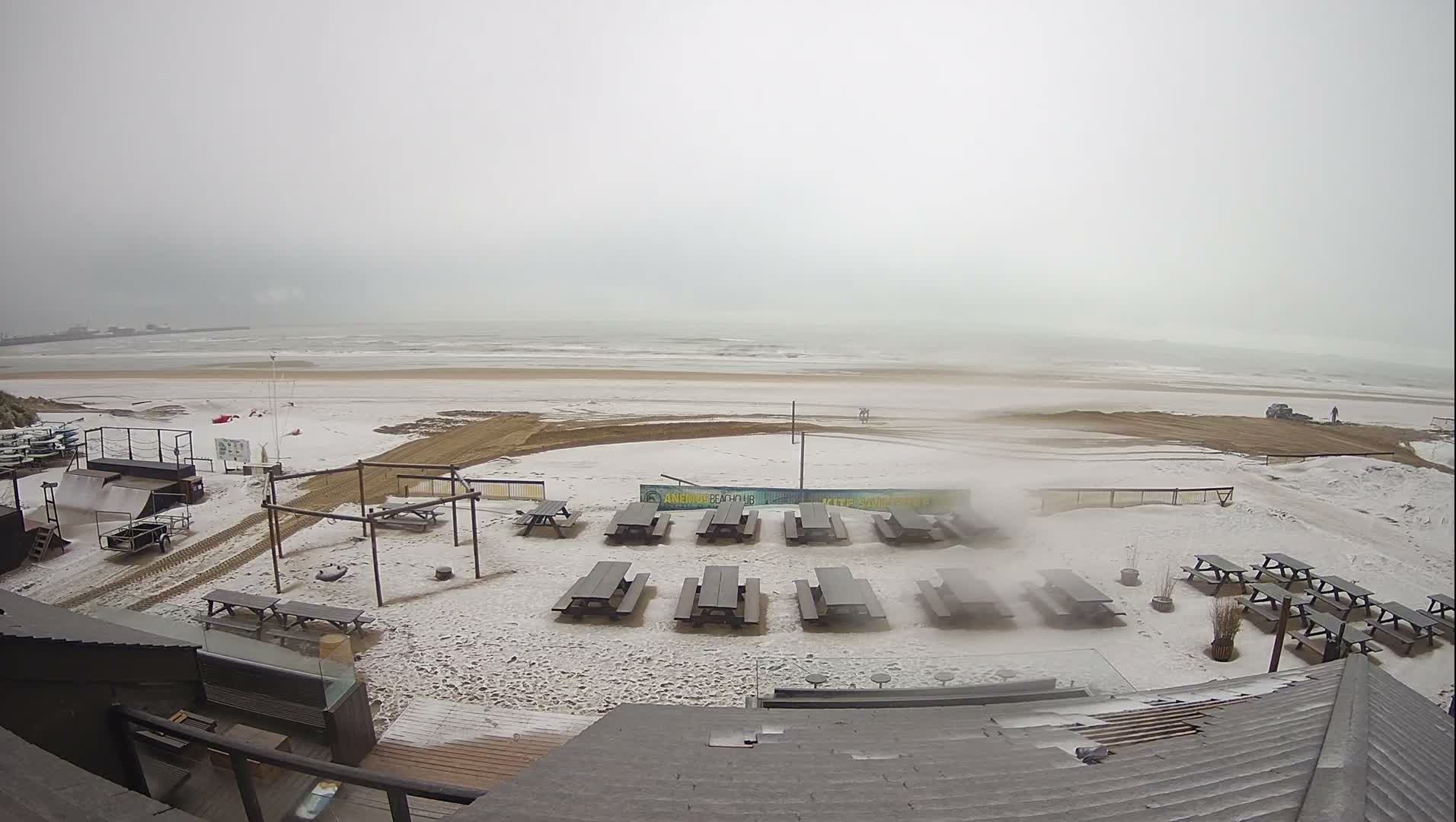 A wide outdoor view captures a snow-covered beach dotted with picnic tables and various beach club structures, extending to a choppy grey ocean beneath a heavily overcast sky.