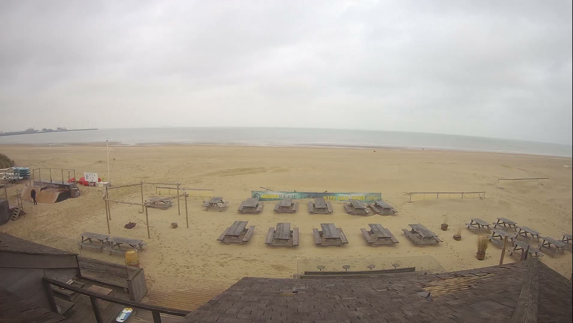 A wide aerial view shows a sandy beach populated with numerous wooden picnic tables and various recreational equipment, leading to an overcast sea with a distant industrial harbor visible under a dull, cloudy sky.