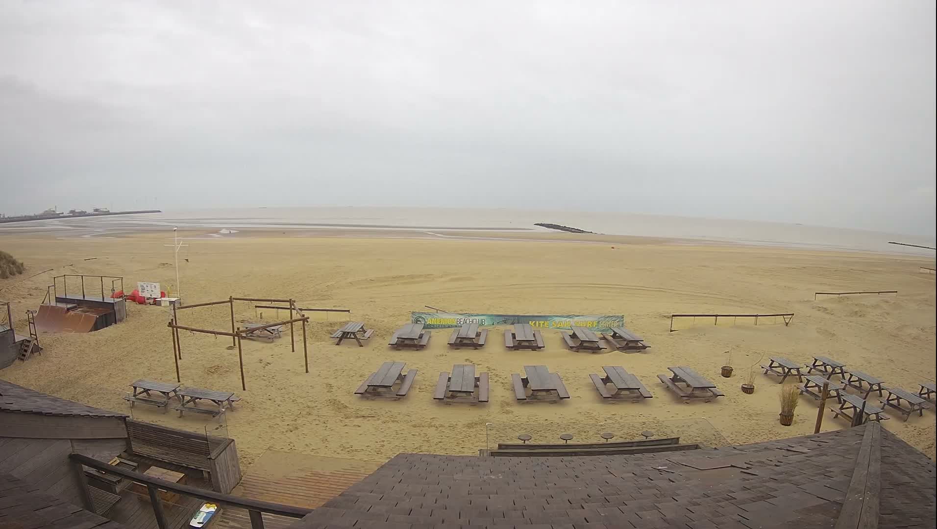 An elevated view captures a wide, sandy beach populated with numerous empty wooden picnic tables, a skateboard ramp, and various other beach structures, all under a uniformly overcast sky with the calm sea stretching to the distant horizon and a visible pier.