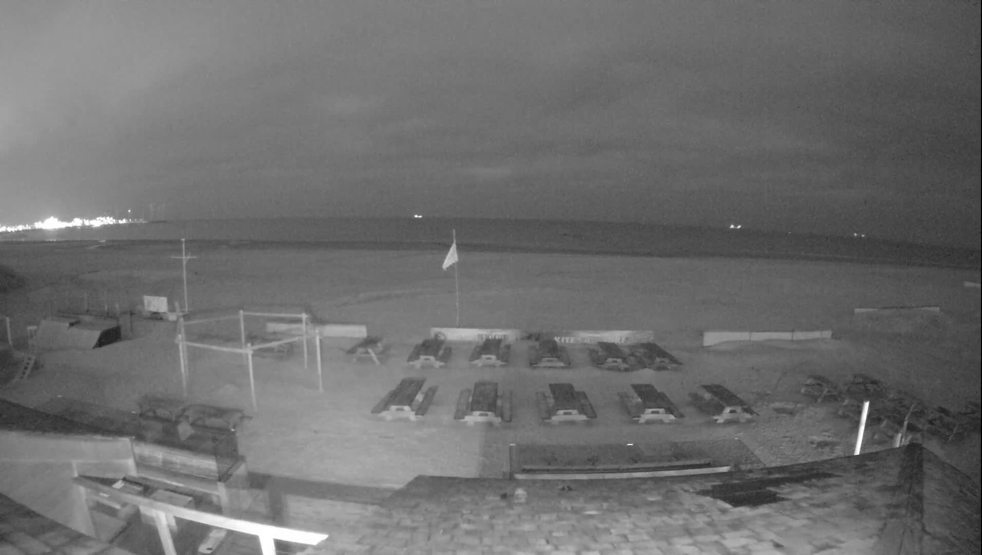 An elevated view captures a wide, sandy beach populated with numerous empty wooden picnic tables, a skateboard ramp, and various other beach structures, all under a uniformly overcast sky with the calm sea stretching to the distant horizon and a visible pier.