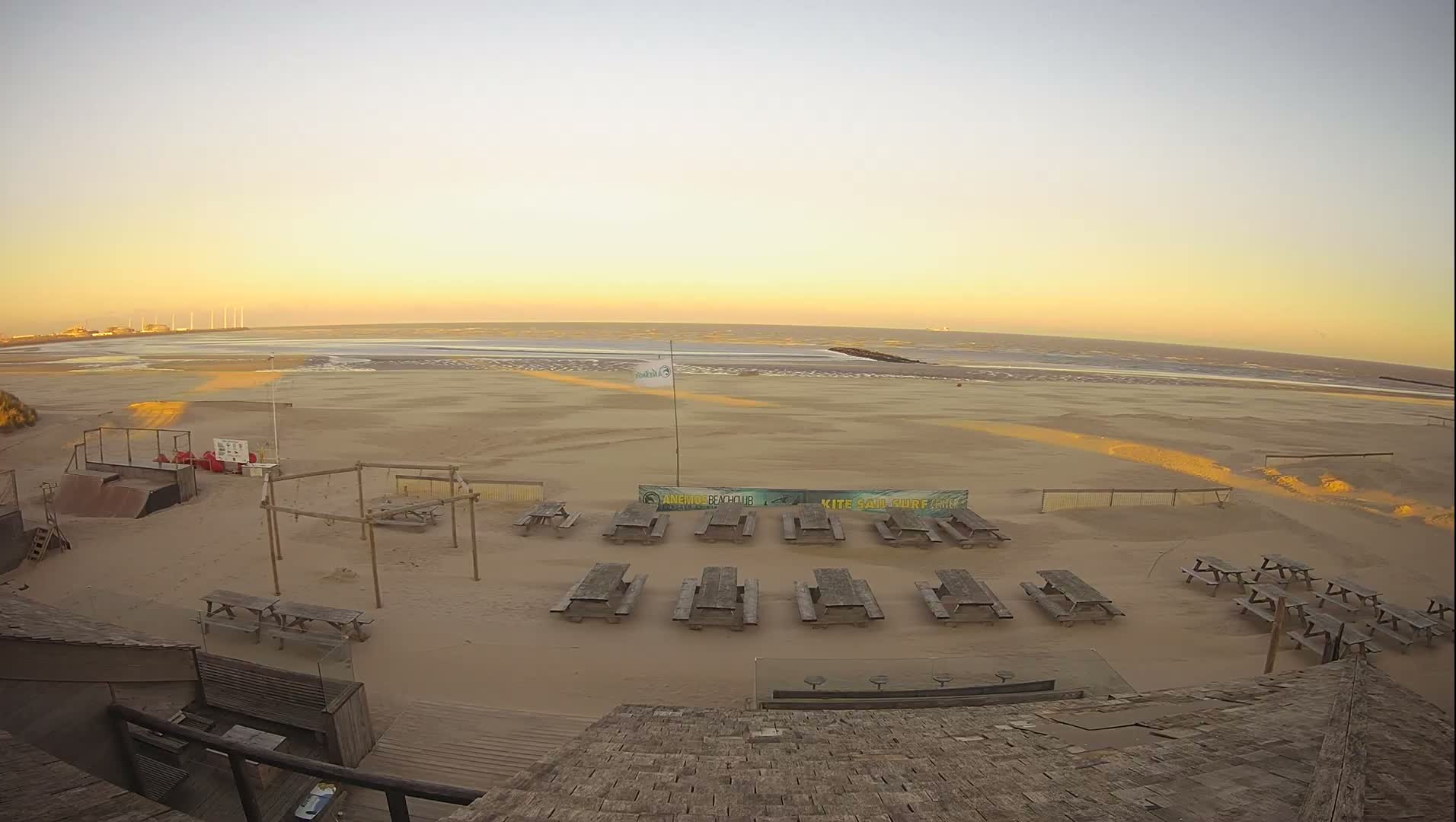 A wide sandy beach featuring numerous wooden picnic tables and structures is bathed in the warm, clear light of a sunrise or sunset, with the calm ocean stretching to the horizon where distant industrial structures are visible.