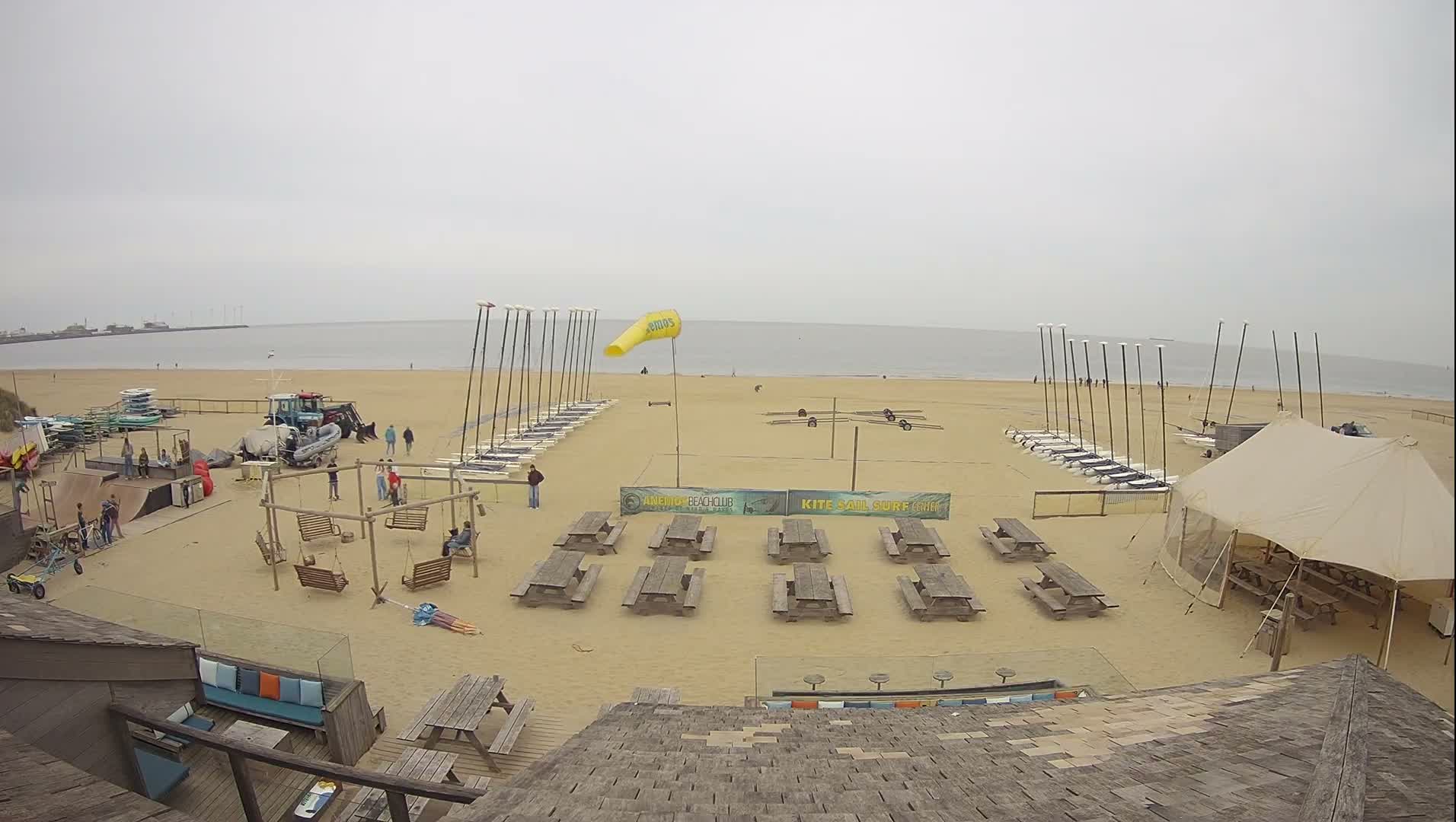A wide sandy beach featuring numerous wooden picnic tables and structures is bathed in the warm, clear light of a sunrise or sunset, with the calm ocean stretching to the horizon where distant industrial structures are visible.