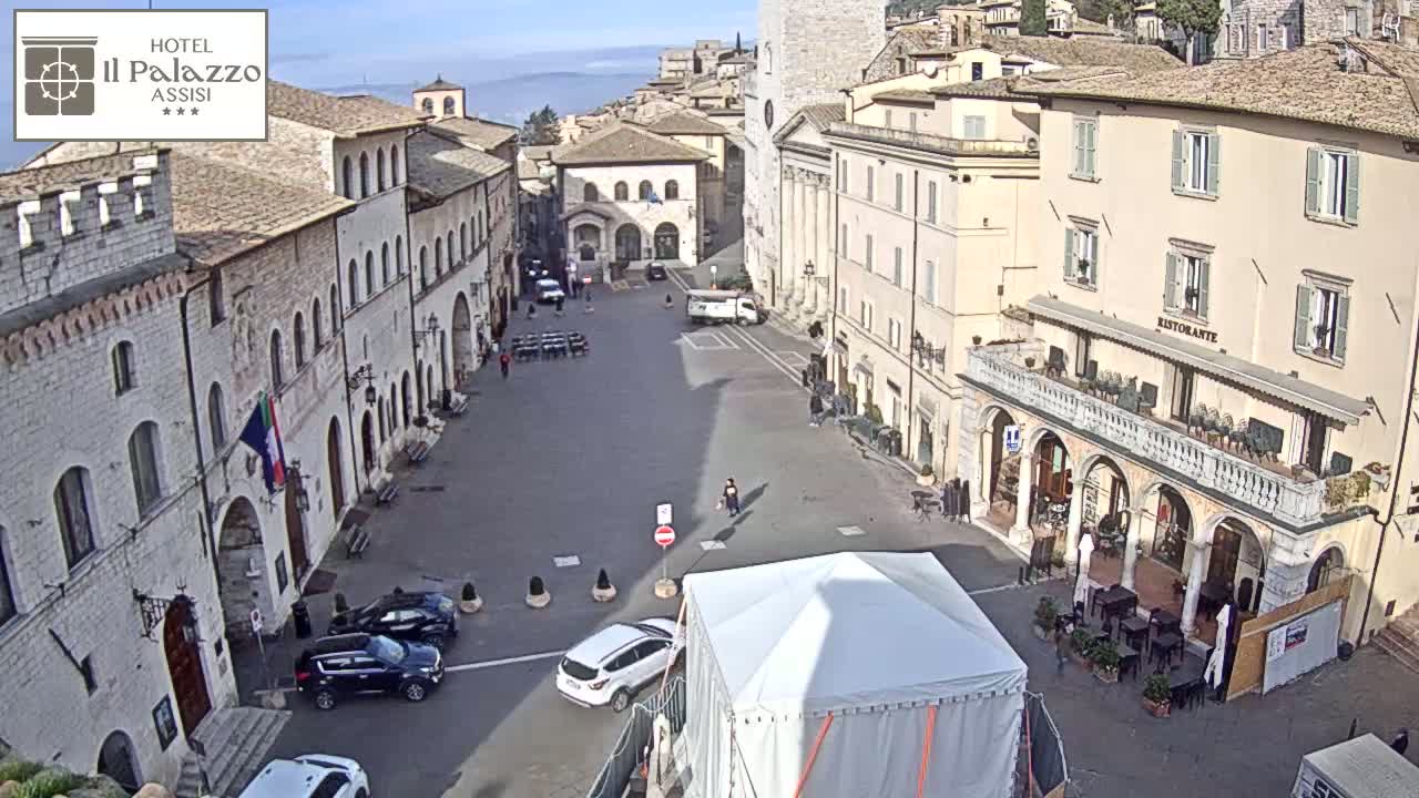 A mostly empty town square in Assisi, Italy, is surrounded by aged stone buildings under a clear, sunny sky.