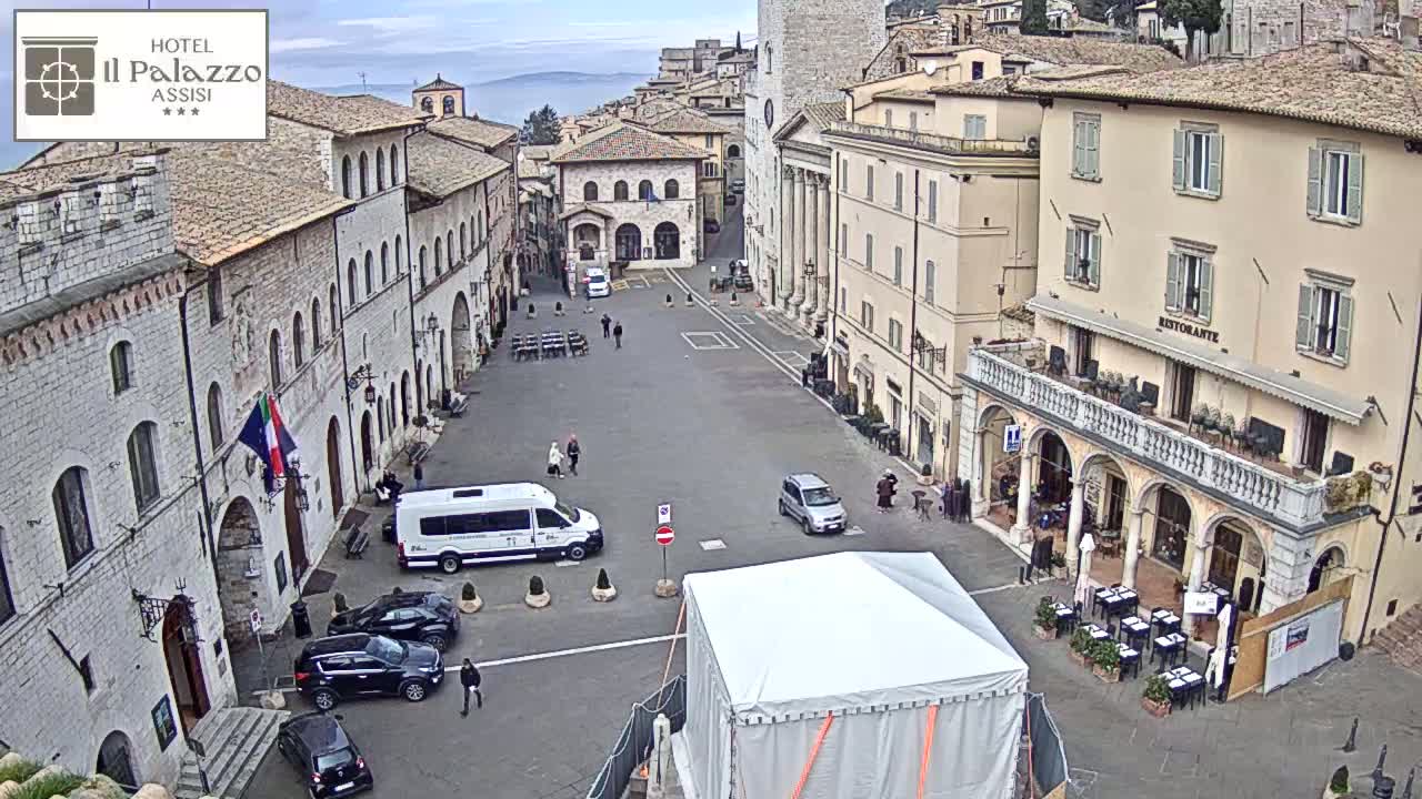 A mostly empty town square in Assisi, Italy, is viewed from above on an overcast day, with several cars parked and a few pedestrians walking around.