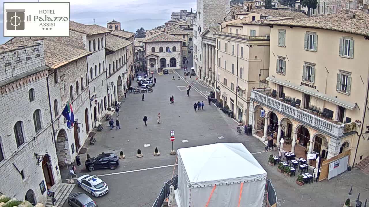 A mostly empty town square in overcast weather is surrounded by ancient-looking buildings, with a few pedestrians and parked cars visible.