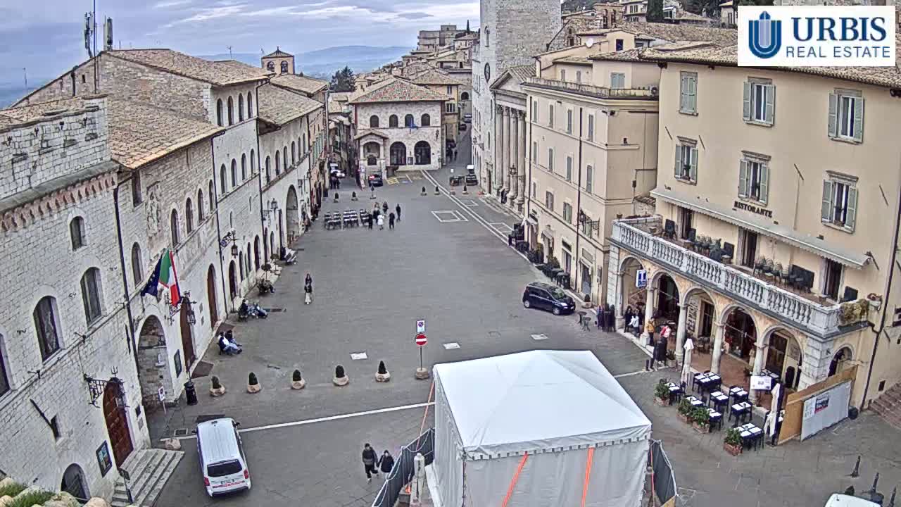 A mostly empty town square in a historic Italian city under a cloudy sky is surrounded by stone buildings, with a few people and cars visible.