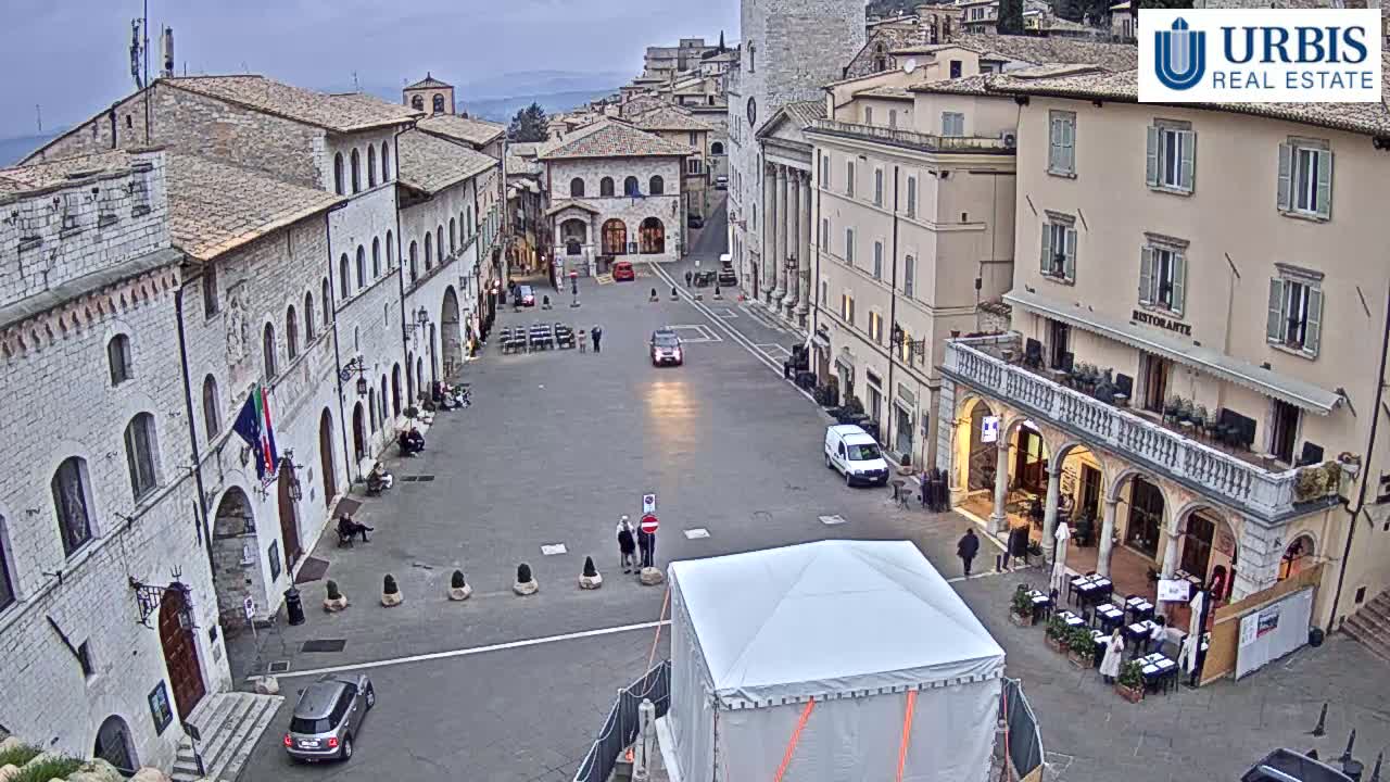 A large, mostly empty town square is surrounded by stone buildings under an overcast sky, with a few cars and pedestrians present.