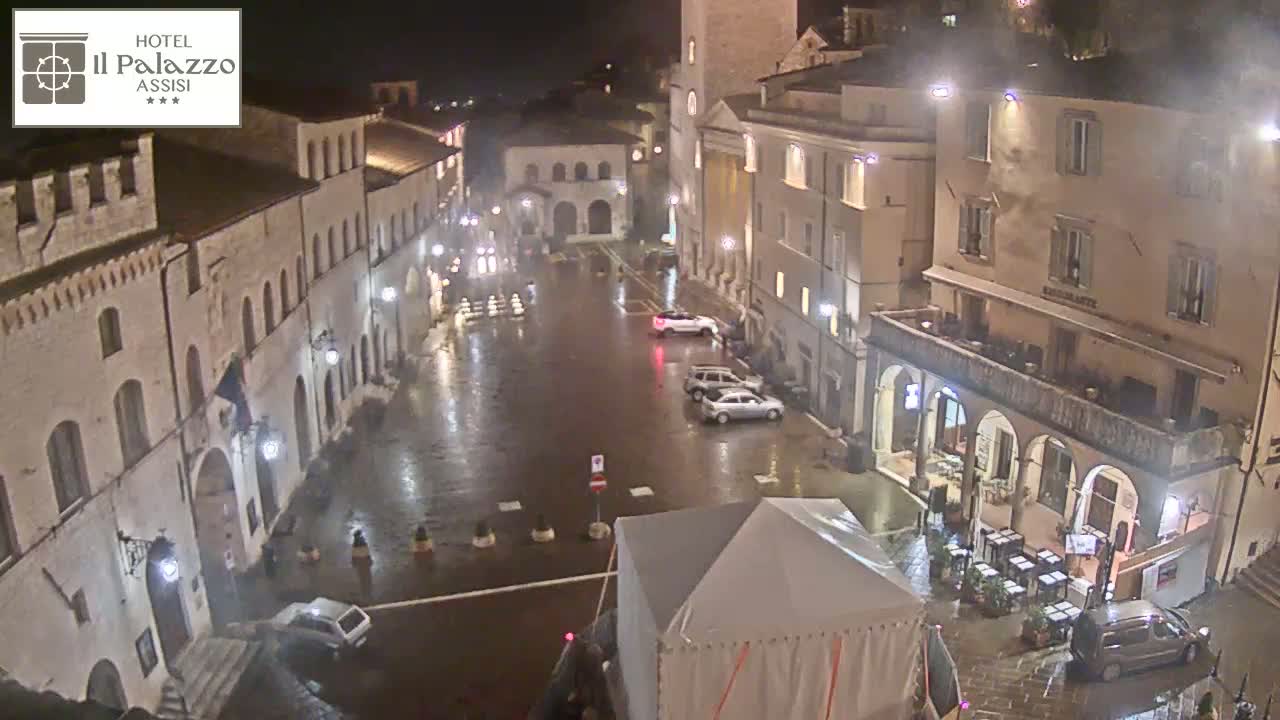 A rain-slicked town square at night, surrounded by old stone buildings, with several cars parked and a few people visible.