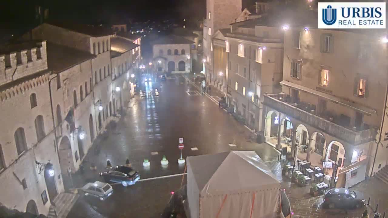 A rain-slicked town square at night, surrounded by illuminated buildings, with a few cars parked and a covered structure in the center.