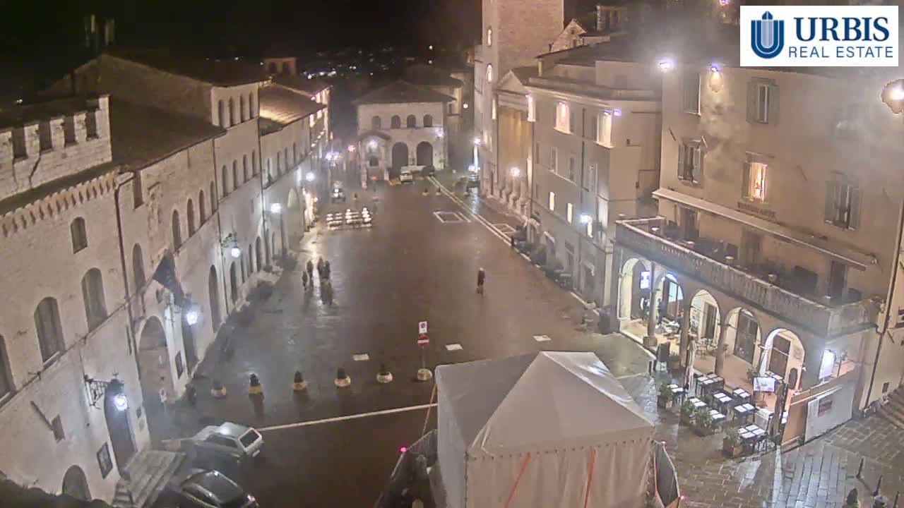 A wet, empty town square at night, surrounded by old stone buildings with arched doorways and windows, is illuminated by streetlights.