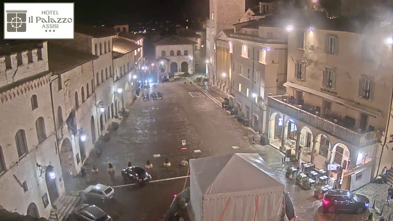 A wet, empty town square at night, surrounded by illuminated buildings, with a few cars parked and a large tent in the center.