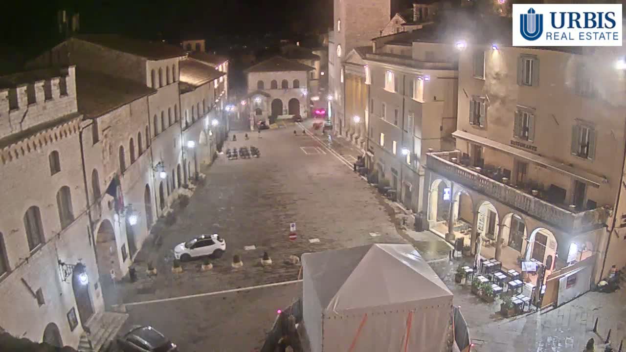 A nighttime, aerial view of a town square surrounded by historical buildings, with a few cars and a temporary tent visible in the mostly empty space.