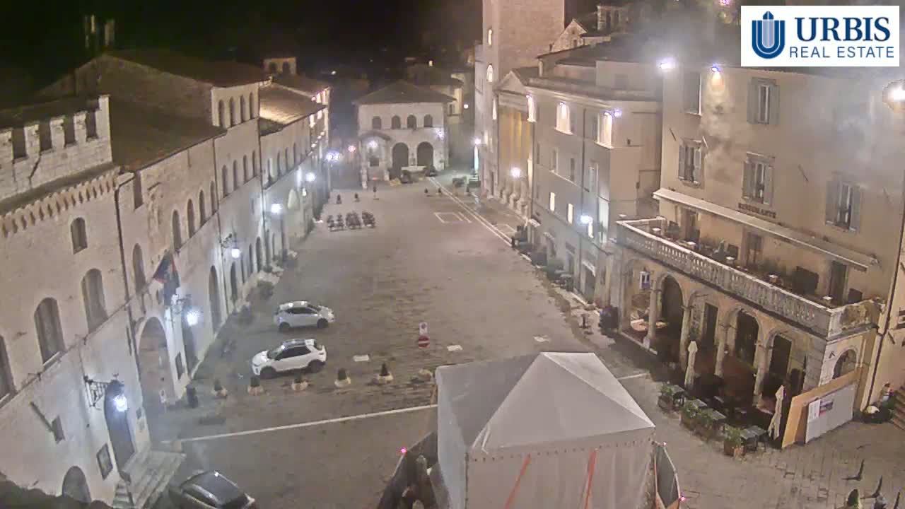 A nighttime view of a mostly empty town square surrounded by historic-looking buildings, illuminated by streetlights.