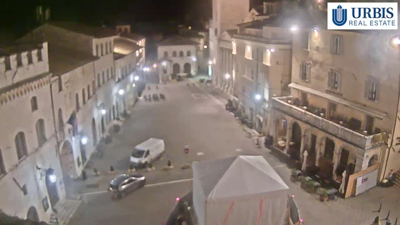 A nighttime, high-angle view of a mostly empty town square surrounded by old, light-colored buildings, with a few vehicles and people visible.