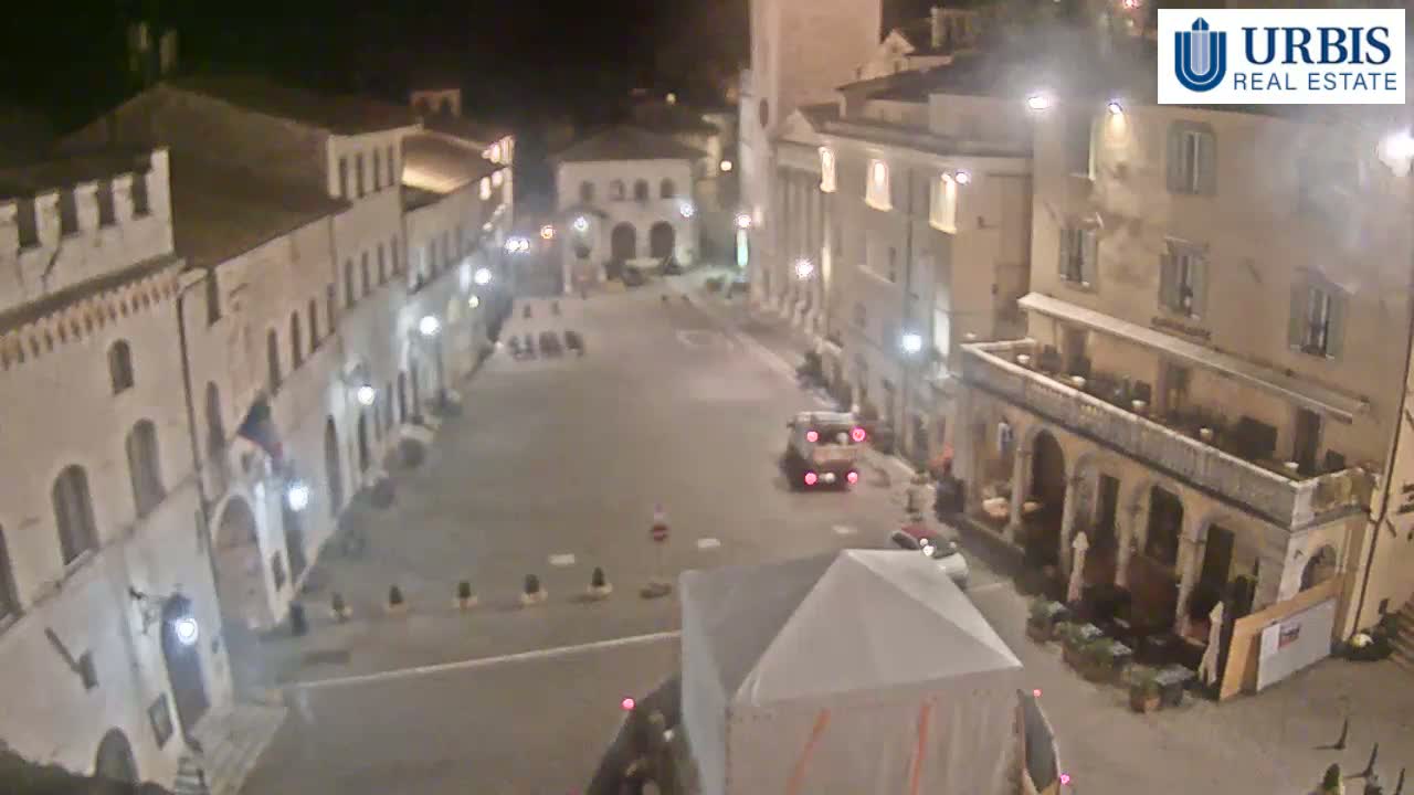 A mostly empty town square at night, surrounded by old, light-colored buildings, is illuminated by streetlights.