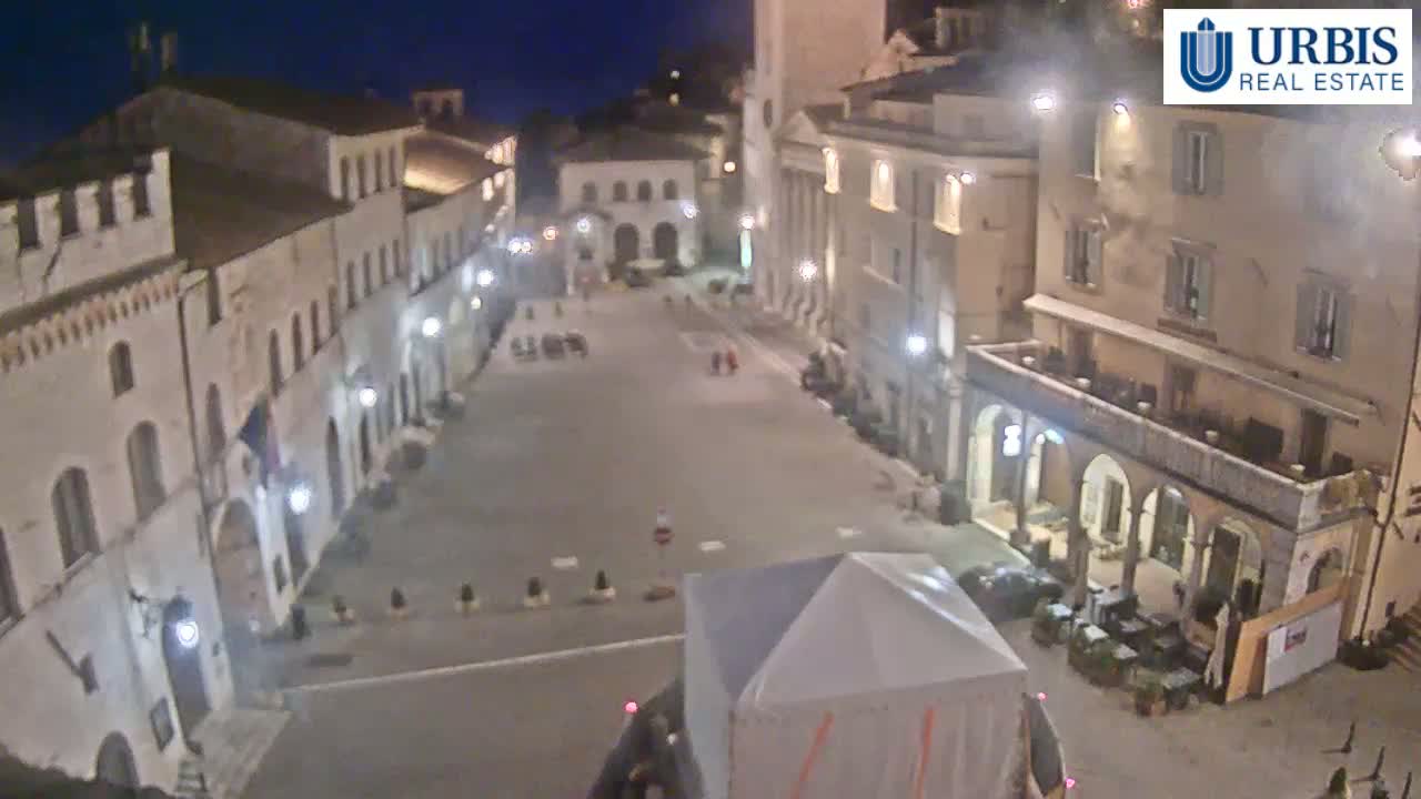 An empty town square at night, surrounded by illuminated buildings, with a white tent in the foreground.