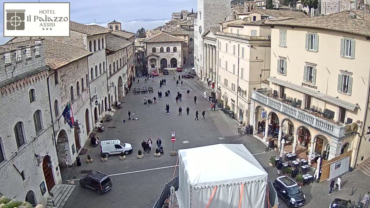 A large, open square in a European town, surrounded by old stone buildings, under a mostly cloudy sky, with a few people and cars scattered about.