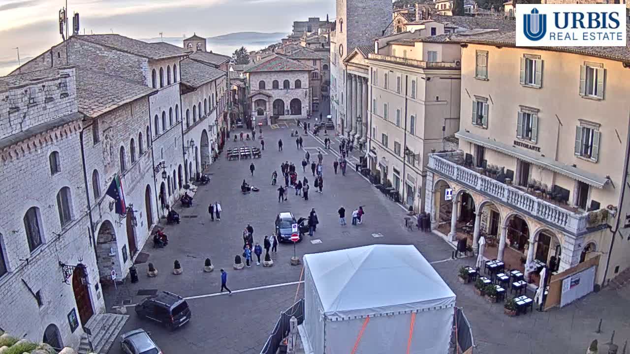 A town square in a historic, stone building-lined city is populated with a few people, cars, and a large white tent, under a mostly clear, but somewhat hazy sky.
