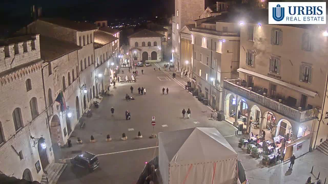 A nighttime, high-angle view of a sparsely populated town square surrounded by historic stone buildings, with a few people walking and sitting at outdoor cafes.