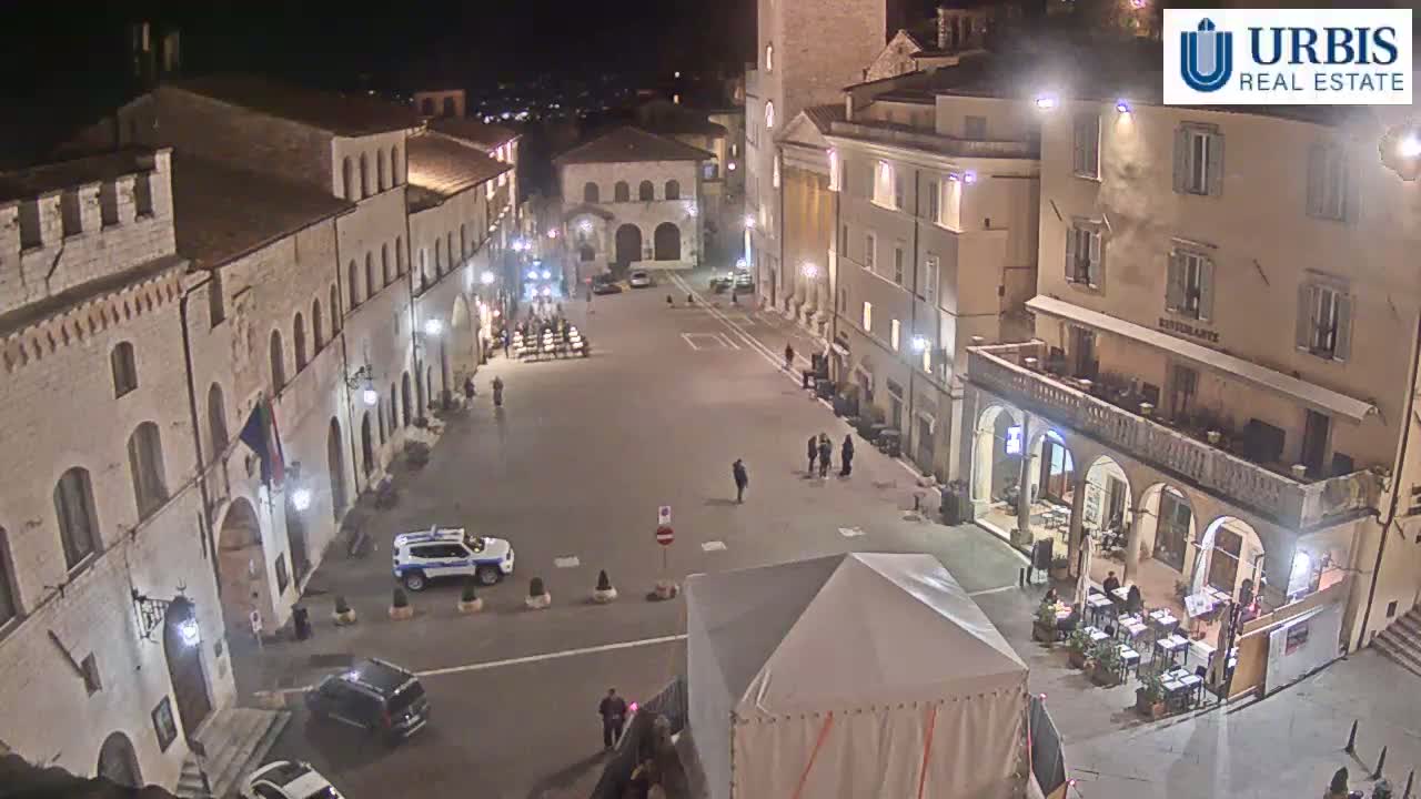 A nighttime, high-angle view of a town square with several buildings, a few cars, and people, lit by streetlights.