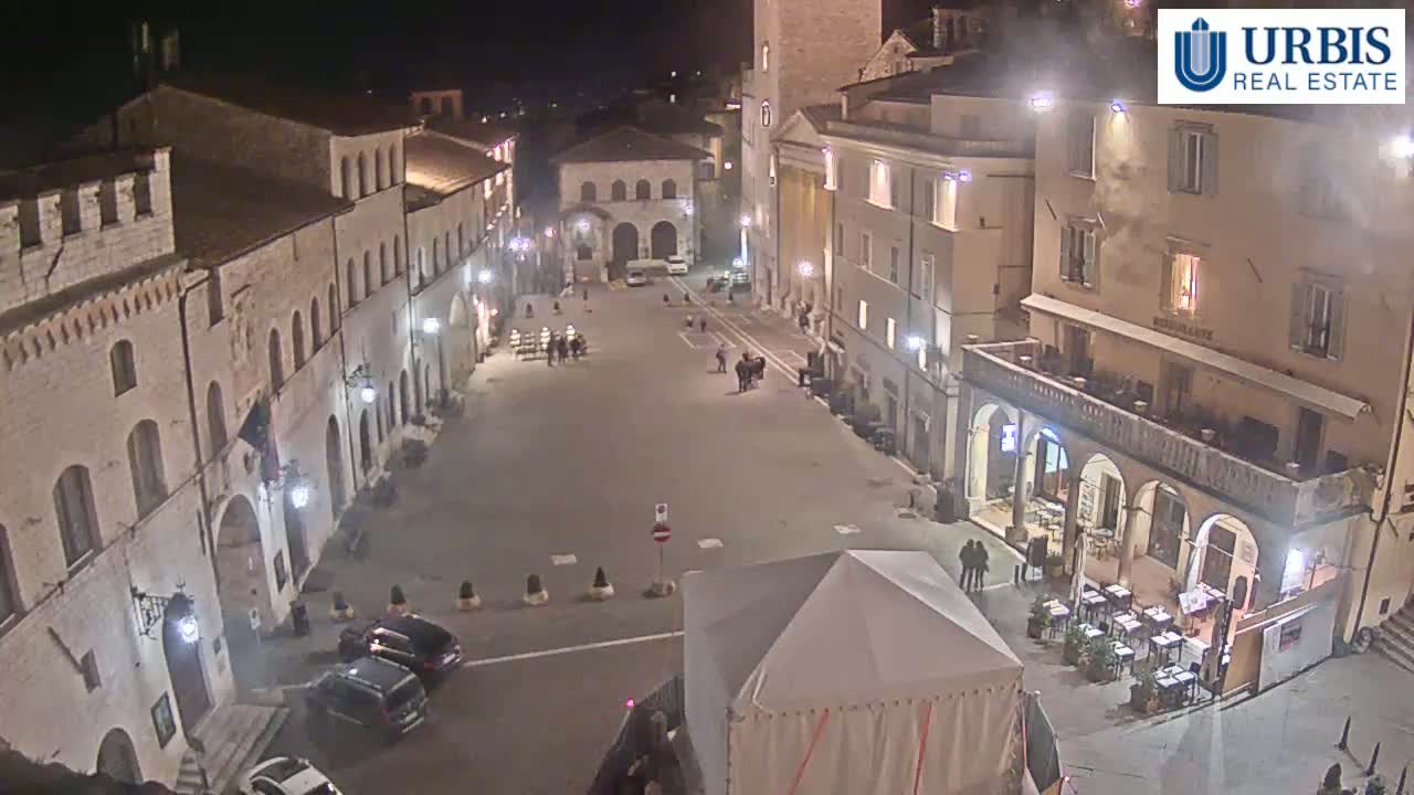 A mostly empty town square at night, illuminated by streetlights, is surrounded by old stone buildings, with a few people and cars visible.