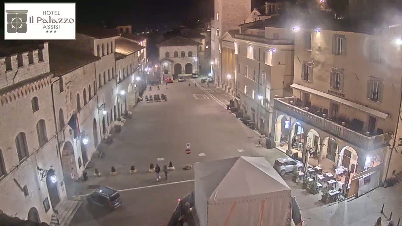 A nighttime, high-angle view of a mostly empty town square surrounded by historic buildings, with a few people and vehicles present.