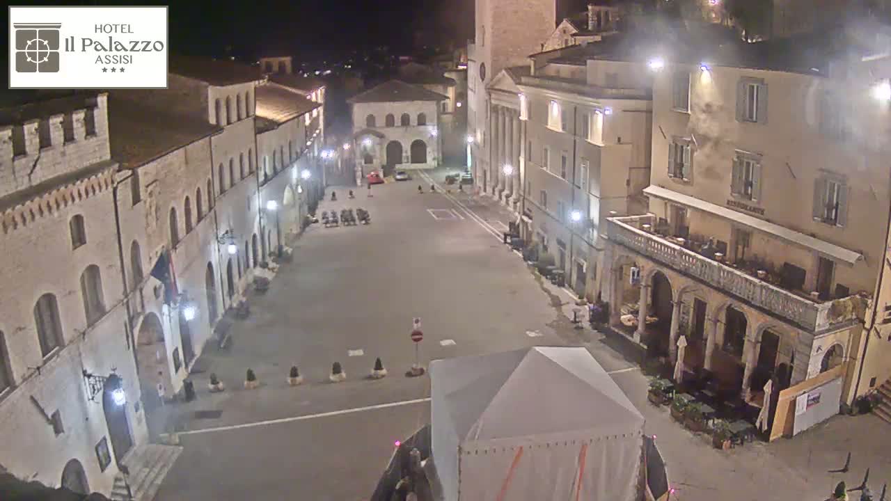 A nighttime view of a sparsely populated town square surrounded by historic-looking buildings under dark, clear skies.