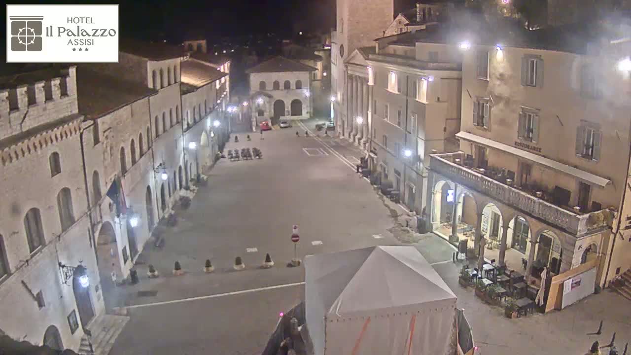 A mostly empty, stone-walled town square at night, illuminated by streetlights, with a few cars and some outdoor seating at a restaurant.