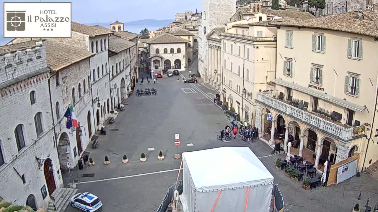 A mostly empty town square in a city with pale stone buildings, under a partly cloudy sky, with a few people and bicycles visible.