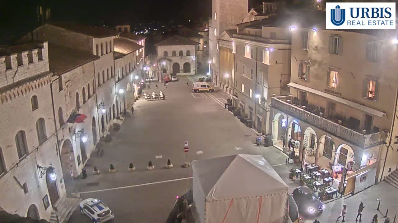 A nighttime, high-angle view of a town square surrounded by historic stone buildings, with a few vehicles and pedestrians present.